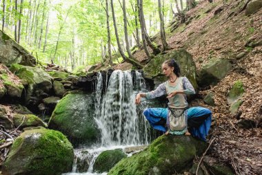 Mavi elbiseli esmer kadın bir dağ şelalesinin yanında oturuyor, Karpat yaz ormanı, Ukrain.