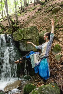 Mavi elbiseli esmer kadın şelalenin yanında dans ediyor, yaz, karpatyalı.