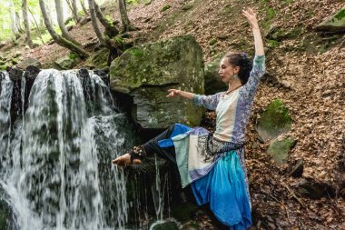 Mavi elbiseli esmer kadın şelalenin yanında dans ediyor, yaz, karpatyalı.