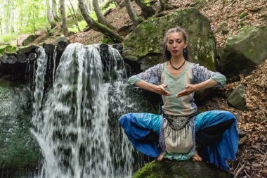 Mavi elbiseli esmer kadın bir dağ şelalesinin yanında oturuyor, Karpat yaz ormanı, Ukrain.