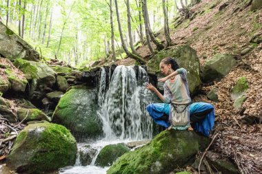 Mavi elbiseli esmer kadın bir dağ şelalesinin yanında oturuyor, Karpat yaz ormanı, Ukrain.