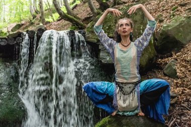 Mavi elbiseli esmer kadın bir dağ şelalesinin yanında oturuyor, Karpat yaz ormanı, Ukrain.