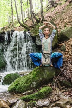 Mavi elbiseli esmer kadın bir dağ şelalesinin yanında oturuyor, Karpat yaz ormanı, Ukrain.