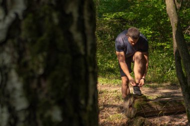 Sporcu, bacaklarının bağcıklarını spor ayakkabılarla bağlıyor. Parkta koşan sporcu, açık havada, ormanın etrafında koşuyor. Meşe ağaçları genç çimenler atletik atletizm ormanı sporcularına dayanıyor.