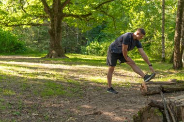 Genç, dayanıklı bir atlet ormanda, ormanın etrafında, meşe ağaçları etrafında esneme hareketleri yapıyor. Sağlıklı orman, yaşam tarzı erkek, dışarıda orman. Sonbahar haçı, ayaklar uzar