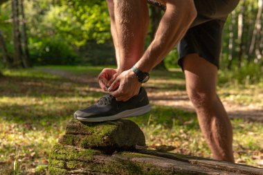 Sporcu, bacaklarının bağcıklarını spor ayakkabılarla bağlıyor. Parkta koşan sporcu, açık havada, ormanın etrafında koşuyor. Meşe ağaçları, yeşil çimenler, atletik spor ormanlarında yaşayan genç sporcular.