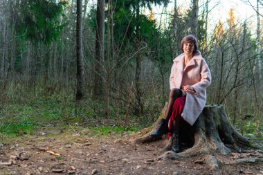 Bir kız, kozalaklı bir ormanda, kırmızı bir elbiseyle pembe bir polto üzerinde kütüğün üzerinde oturuyor.