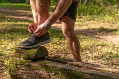 Sporcu, bacaklarının bağcıklarını spor ayakkabılarla bağlıyor. Parkta koşan sporcu, açık havada, ormanın etrafında koşuyor. Meşe ağaçları, yeşil çimenler genç sporcu spor koşusu yapıyor.