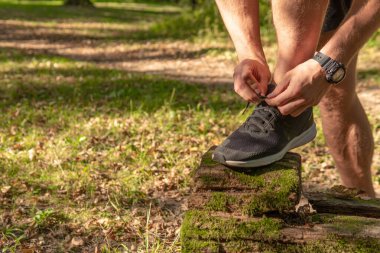 Sporcu, bacaklarının bağcıklarını spor ayakkabılarla bağlıyor. Parkta koşan sporcu, açık havada, ormanın etrafında koşuyor. Meşe ağaçları, yeşil çimenler genç sporcu koşucu doğası gereği koşuyor.