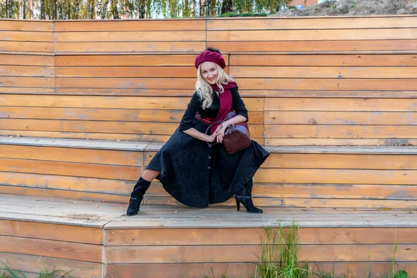 Una donna gioiosa siede su una panchina del teatro estivo, fatta di legno in cappotto bordeaux e biret, nella caduta contro il cielo blu. — Foto Stock