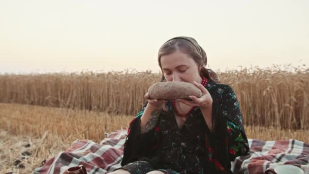 Uma menina em um campo de trigo quebra um pão e cheira o cheiro de pão. — Vídeo de Stock