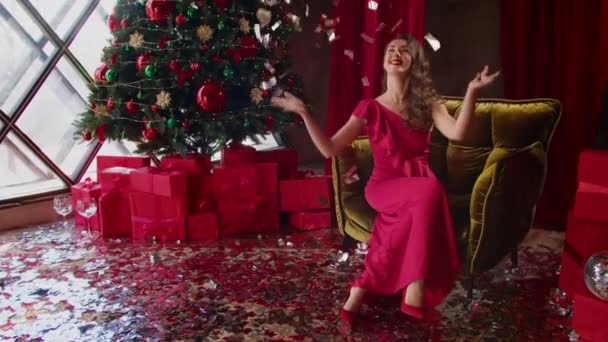 Smuk glad brunette kvinde i rød aftenkjole. kvinde griner, spreder skinnende festligt papir. berømtheder. Jul og nytår gaver fra tryllekunstneren og et træ på hotellet. – Stock-video