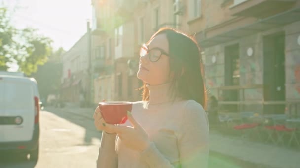 Zrelaksowana uśmiechnięta zadowolona kobieta z czerwoną filiżanką kawy stojąca na końcu ulicy ciesząca się słonecznym dniem — Wideo stockowe