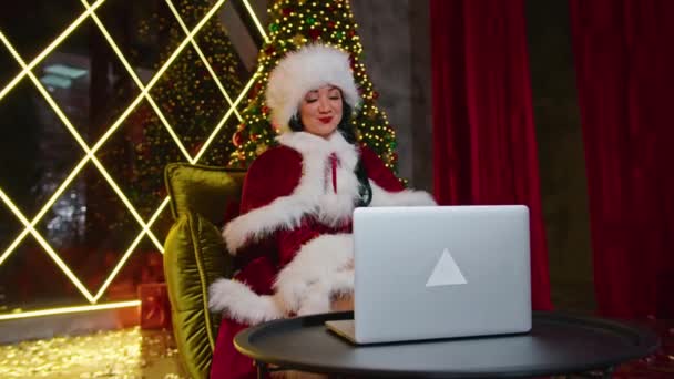 Gelukkige vrouw mevrouw Kerstman communiceert op laptop in haar kerstkantoor. vakantie in quarantaine. communicatie op afstand, vakantiegeschenken en kortingen. — Stockvideo