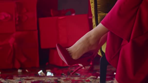Közelkép, egy piros ruhás nő piros cipőt vesz fel, egy fotelban ül egy hotelszobában, egy nő készül a karácsony ünnepére. — Stock videók