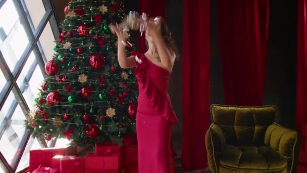 Hermosa mujer morena feliz en vestido de noche rojo. mujer se ríe, dispersando papel festivo brillante. celebridades. Regalos de Navidad y Año Nuevo del mago y un árbol en el hotel. — Vídeo de stock