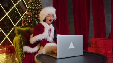 Mutlu kadın Bayan Noel Baba, Noel ofisinde dizüstü bilgisayarla iletişim kuruyor. Karantinada tatiller. Uzaktan iletişim, tatil hediyeleri ve indirimler.