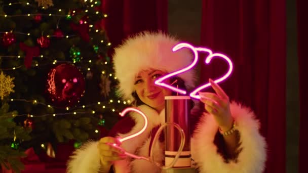 En lycklig kvinna i tomtedräkt leker med neonnummer nära en tesla spole, mot bakgrund av en julgran. Nyårsföreställning, barnledighet. — Stockvideo