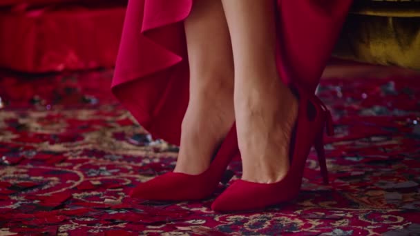 Close-up, seorang wanita dalam gaun merah mengenakan sepatu merah, duduk di kursi di sebuah kamar hotel, seorang wanita sedang mempersiapkan diri untuk liburan Natal. — Stok Video