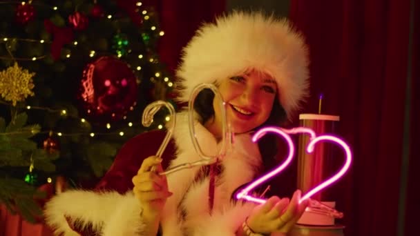 En lycklig kvinna i tomtedräkt leker med neonnummer nära en tesla spole, mot bakgrund av en julgran. Nyårsföreställning, barnledighet. — Stockvideo