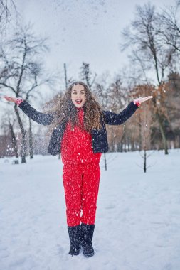 Genç ve mutlu bir kadın kış parkında eğleniyor, kar atıyor, elleri soğuk, salınımlar ölçüsüz..