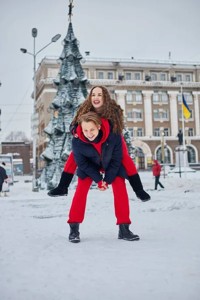 Young family guy and girl spend the day in the park on a snowy day. Emotional young couple having fun while walking in the winter city, a lively man hugs his laughing beautiful woman. — Stock Photo, Image