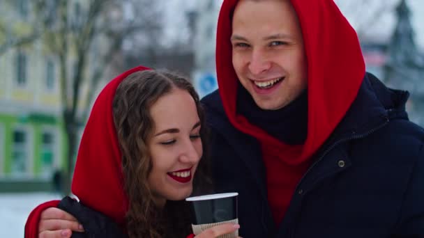 Młody chłopak i dziewczyna spędzają dzień w parku w śnieżny dzień. facet przytula dziewczynę stojąc na ulicy, piją razem kawę. — Wideo stockowe