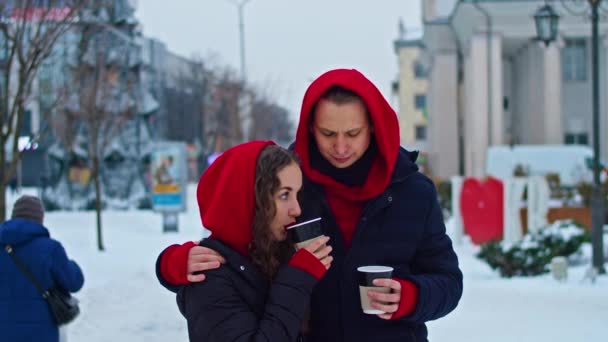 Young family guy and girl spend the day in the park on a snowy day. the guy hugs the girl while standing on the street, they drink coffee together. — Vídeo de Stock