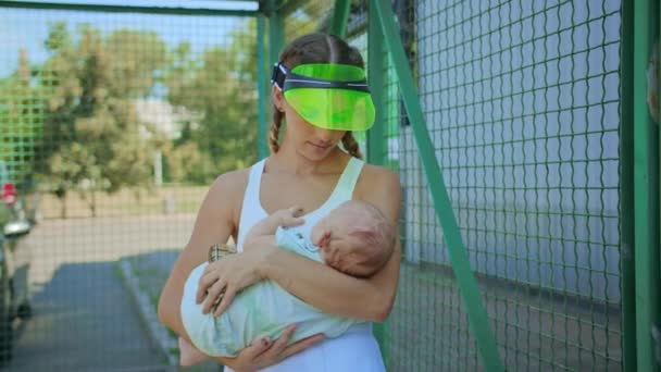 Mujer joven entrenador de tenis con un bebé recién nacido en sus brazos está descansando en la sombra, día soleado caliente. — Vídeos de Stock