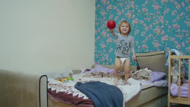 Uma pequena criança feliz pula na cama em casa e joga bola com seu pai, o pai joga a bola para ele fora da tela. — Vídeo de Stock