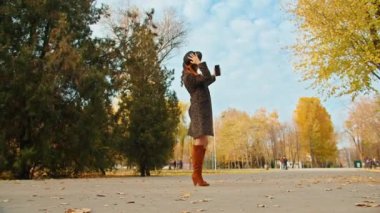 Paltolu, şapkalı ve eşarplı mutlu bir kadın sonbahar parkında telefonda fotoğraf çekerken kendini çekiyor..