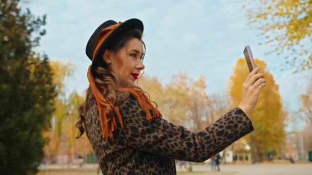 Feliz mulher brincalhão de casaco, chapéu e cachecol no parque de outono filmando-se no telefone tirar fotos. — Vídeo de Stock