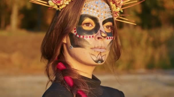 Egy fiatal nő Santa Muerte smink öltözött fekete ruhát a halál sétál a háttérben őszi levelek az erdőben naplementekor. Day of the Dead vagy Halloween koncepció. — Stock videók
