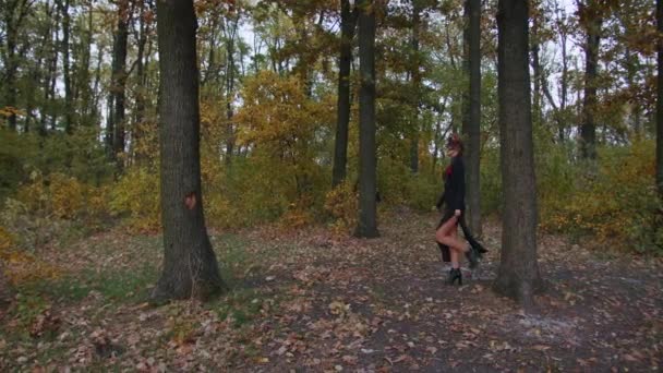 Santa Muerte makyajlı, siyah bir ölüm elbisesi giymiş genç bir kadın günbatımında ormanda sonbahar yapraklarının arka planına doğru yürüyor. Ölüler Günü veya Cadılar Bayramı konsepti. — Stok video