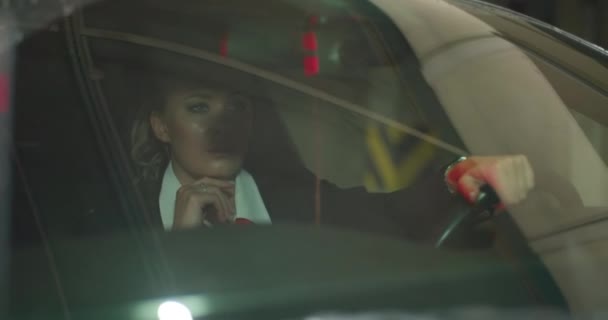 Una hermosa mujer rubia en un traje negro, camisa blanca y corbata roja se sienta en su coche negro en un estacionamiento subterráneo, mirando severamente a un lado. marco cinematográfico, cámara lenta. — Vídeo de stock