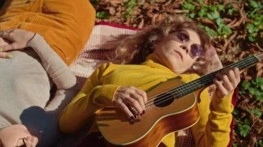 İki mutlu genç kadın şehir parkında harika vakit geçiriyor, bir kız gitar çalıyor, ikincisi ise müziğin ve ağaçların gürültüsünün tadını çıkarıyor. sonbahar yaz dönemi.
