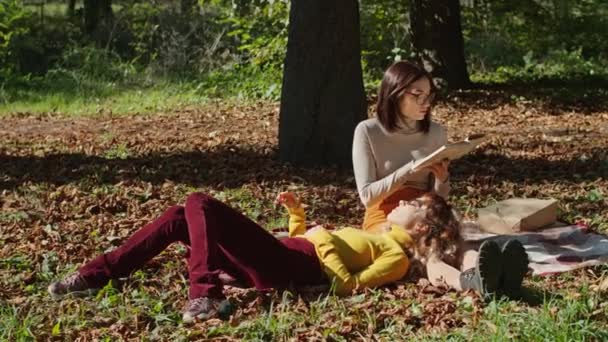 Két boldog fiatal nő jól érzi magát a városi parkban ülve. Boldog fiatal életet. Az egyik lány könyvet olvas, a másik a lábán fekszik.. — Stock videók