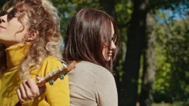 İki mutlu genç kadın şehir parkında harika vakit geçiriyor, bir kız gitar çalıyor, ikincisi ise müziğin ve ağaçların gürültüsünün tadını çıkarıyor. sonbahar yaz dönemi.