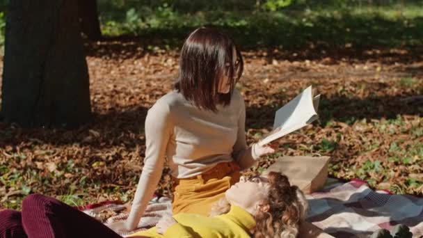 Δύο χαρούμενες νεαρές γυναίκες διασκεδάζουν στο πάρκο της πόλης. Ευτυχισμένη νέα ζωή. Ένα κορίτσι διαβάζει ένα βιβλίο, το δεύτερο βρίσκεται στα πόδια της. — Αρχείο Βίντεο