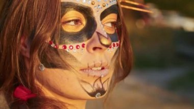 Santa Muerte makyajlı, siyah bir ölüm elbisesi giymiş genç bir kadın günbatımında ormanda sonbahar yapraklarının arka planına doğru yürüyor. Ölüler Günü veya Cadılar Bayramı konsepti.