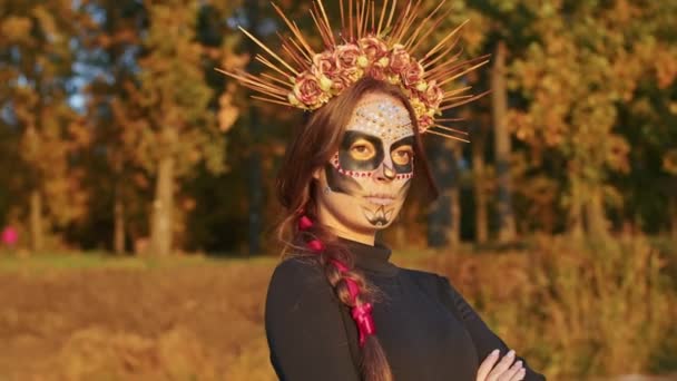 Seorang wanita muda dengan tata rias Santa Muerte mengenakan gaun hitam kematian berjalan dengan latar belakang daun musim gugur di hutan saat matahari terbenam. Konsep Hari Kematian atau Halloween. — Stok Video