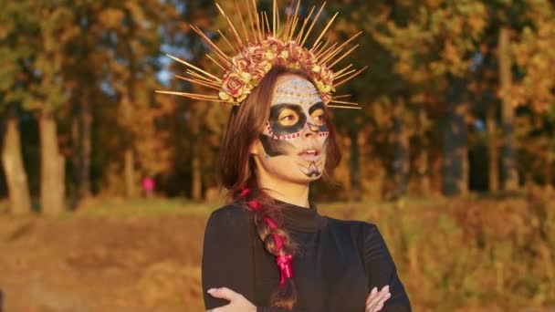 Egy fiatal nő Santa Muerte smink öltözött fekete ruhát a halál sétál a háttérben őszi levelek az erdőben naplementekor. Day of the Dead vagy Halloween koncepció. — Stock videók