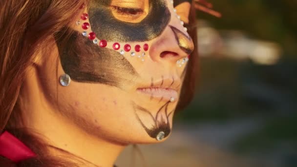 Eine junge Frau mit Santa-Muerte-Make-up in einem schwarzen Totenkleid spaziert bei Sonnenuntergang vor dem Hintergrund von Herbstblättern durch den Wald. Totensonntag oder Halloween-Konzept. — Stockvideo