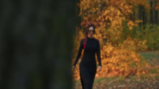 Молодая женщина с макияжем Санта-Муэрте, одетая в черное платье смерти, идет на фоне осенних листьев в лесу на закате. День мертвых, или Хэллоуин. — стоковое видео