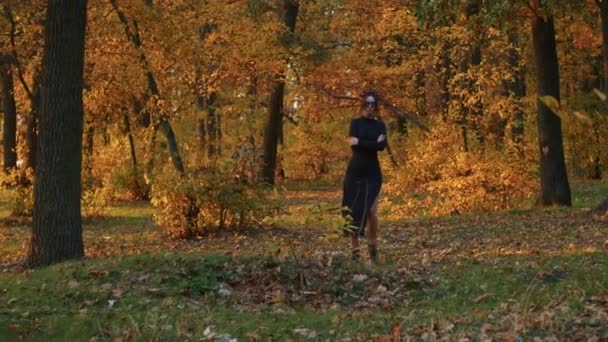 Een jonge vrouw met Santa Muerte make-up gekleed in een zwarte jurk des doods wandelt tegen de achtergrond van herfstbladeren in het bos bij zonsondergang. Dag van de Dood of Halloween concept. — Stockvideo