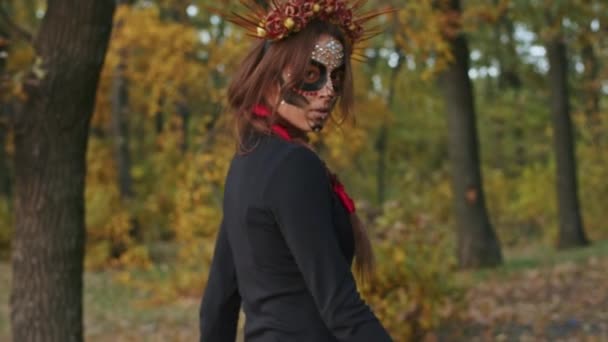 Uma jovem mulher com maquiagem Santa Muerte vestida com um vestido preto da morte caminha contra o pano de fundo das folhas de outono na floresta durante o pôr do sol. Conceito Dia dos Mortos ou Halloween. — Vídeo de Stock