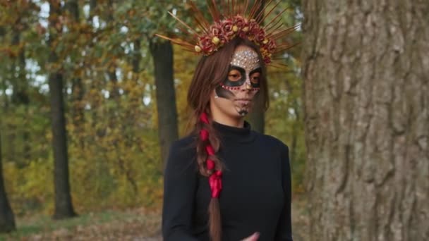 Uma jovem mulher com maquiagem Santa Muerte vestida com um vestido preto da morte caminha contra o pano de fundo das folhas de outono na floresta durante o pôr do sol. Conceito Dia dos Mortos ou Halloween. — Vídeo de Stock