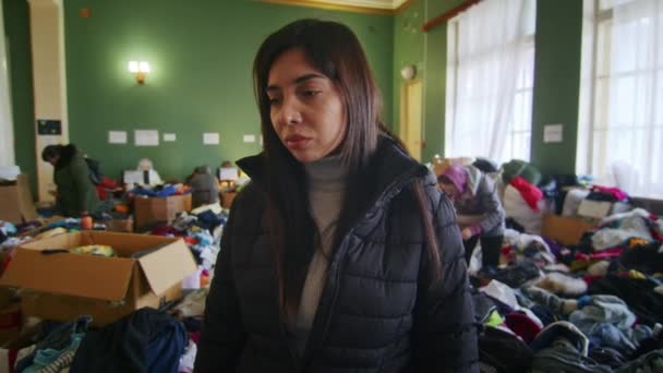 Ett porträtt av en ledsen armenisk kvinna i den stora salen på flyktingförläggningen bland ett stort antal kläder och flyktingar. Krig mellan Ryssland och Ukraina. — Stockvideo
