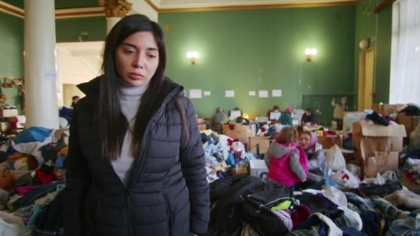 Un retrato de una mujer armenia triste en el gran salón del centro de asistencia a refugiados entre un gran número de ropa y refugiados. Guerra entre Rusia y Ucrania. — Vídeos de Stock