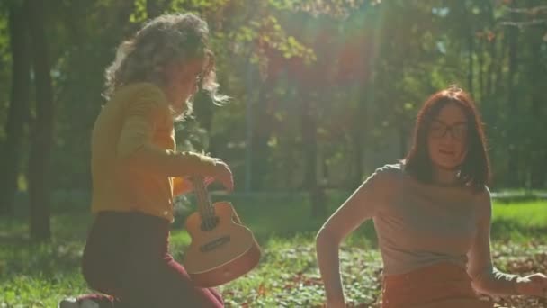 Dos jóvenes felices se están divirtiendo en el parque de la ciudad, una chica está tocando la guitarra, la segunda está disfrutando de la música y el ruido de los árboles. otoño período de verano. — Vídeos de Stock